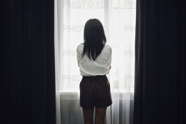 A woman stands with her back to the viewer, gazing out of a bright window. The soft afternoon light