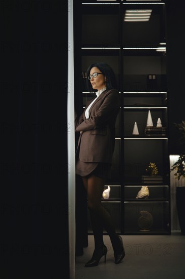A confident woman in brown jacket, mini skirt and glasses stands with her arms crossed in an office