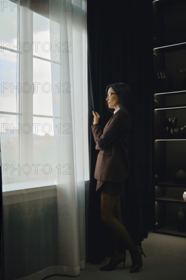 A woman dressed in a tailored suit stands by a large window, pulling back sheer curtains. She