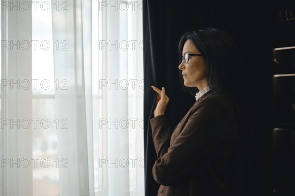 A businesswoman stands by a window in a modern office, gazing thoughtfully outside. She wears