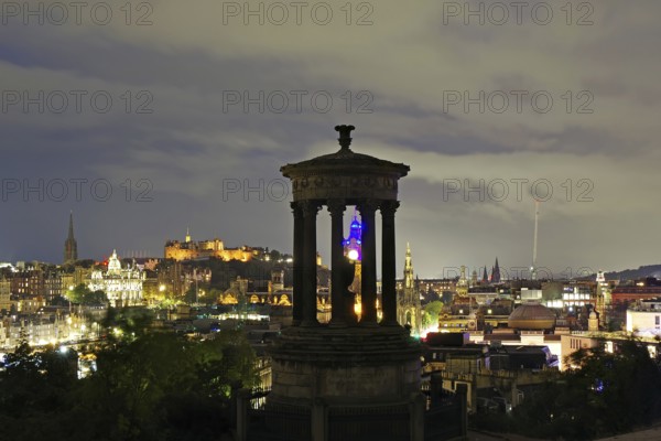Night scene of Edinburgh with illuminated buildings and a historical monument in the foreground,