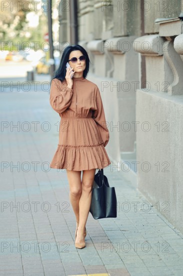 A fashionable woman strolls down the city sidewalk wearing a brown dress and sunglasses, carrying a