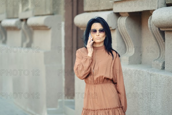 A woman wearing a brown dress and sunglasses engages in a phone conversation while passing by the