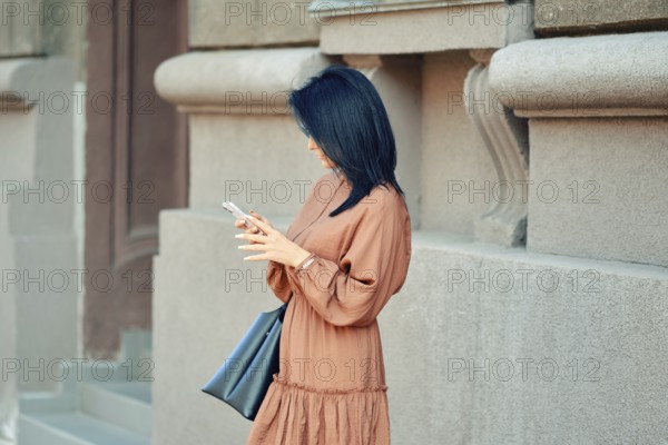A woman in a flowing brown dress and a handbag stands against a stylish urban building. She is