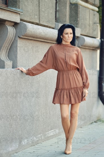 A relaxed woman stands by a textured stone wall, wearing a stylish brown dress with a flowing skirt