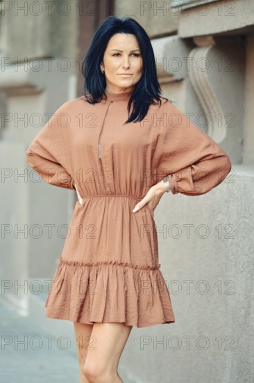 A woman stands with her arms on waist on a city sidewalk, wearing a chic, loose-fitting dress. Her