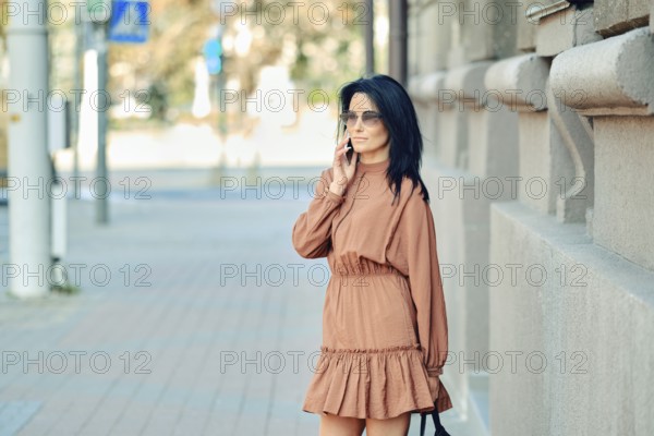 A fashionable woman dressed in a brown dress engages in a phone conversation while standing on a