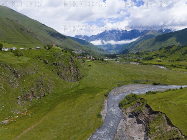 River flowing through green valley, surrounded by mountains and clouds, near a small village,