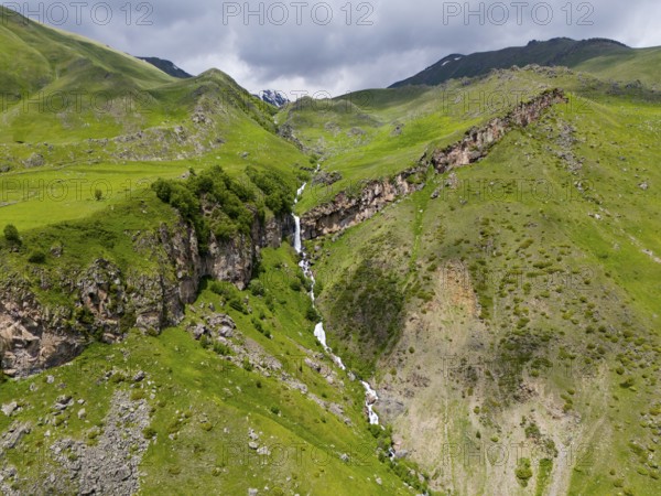 Waterfall cascading down the rocky mountain face, surrounded by green landscape, aerial view, Toti