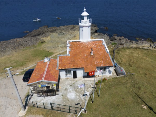 A lighthouse on a rocky coast with blue sky and sea, next to it a building with a terracotta roof,
