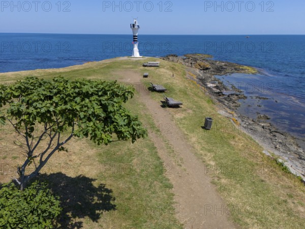 Path leading to a lighthouse on the coast, surrounded by trees and benches in a summer landscape,