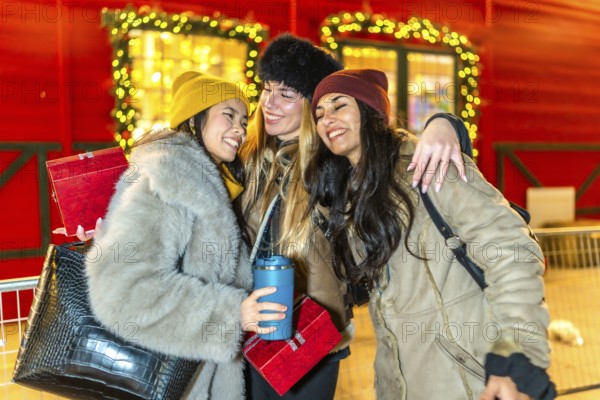Three happy female friends hugging at a decorated christmas market stall at night, holding