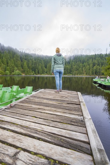 Woman standing alone on a wooden jetty in front of a quiet lake surrounded by forest, Mummelsee,