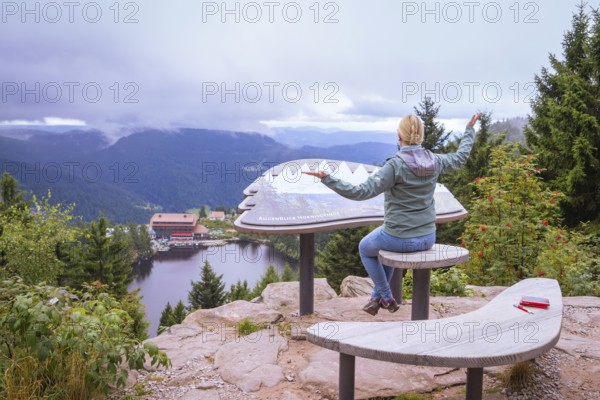 Woman sitting on a wooden bench at a viewpoint, overlooking a lake and mountains, Mummelsee,