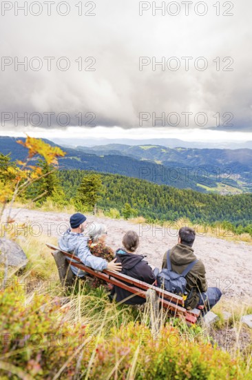 Three people on a bench enjoy the wide view over wooded mountains and valleys, Mummelsee, Seebach,