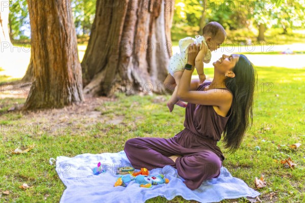 Young latin mother playing with her baby girl in a park, lifting her in the air and smiling,