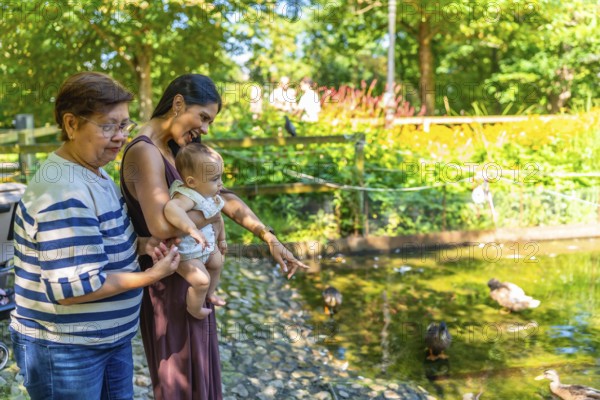 Grandmother, mother, and granddaughter enjoying a sunny day at the park, observing ducks swimming