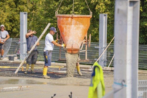 Two workers pouring concrete from large mixer tubs, surrounded by safety measures and mesh fencing,