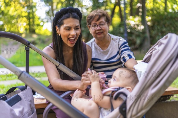 Grandmother, mother and baby girl enjoying a sunny day at the park, feeding the baby sitting in a
