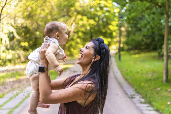 Smiling latin mother lifting her baby daughter while playing together in a sunny park, enjoying a