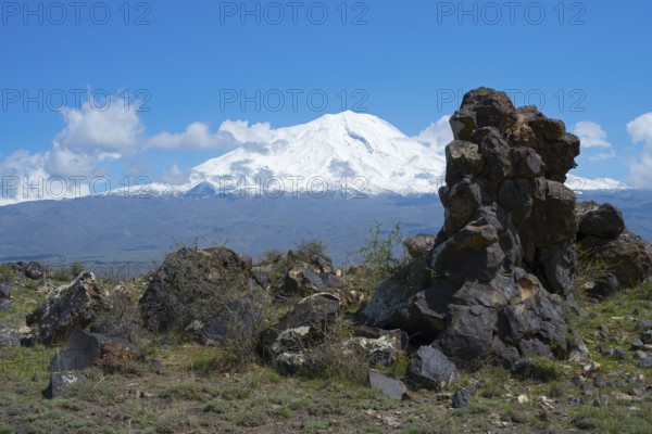 Snow-covered mountain under blue sky with clouds, surrounded by rocks and green landscape, aerial