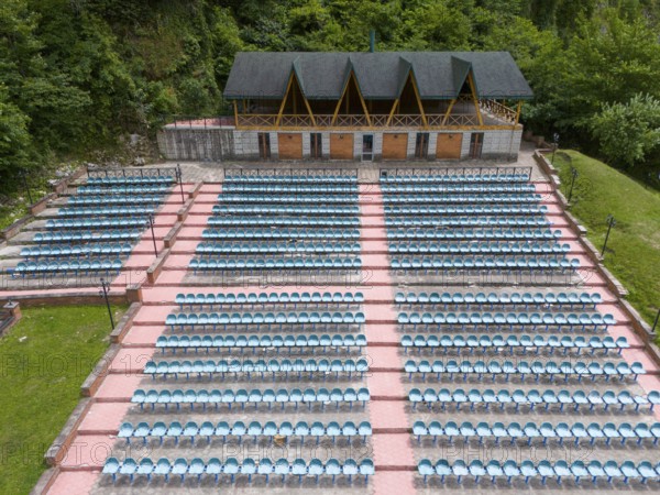 An amphitheatre with many rows of seats and a covered stage against a wooded backdrop, aerial view,