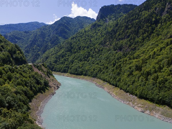 A turquoise-coloured river winds through a wooded and mountainous valley under a clear sky, aerial