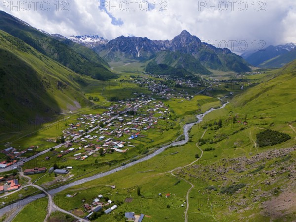 Wide valley with river and village, surrounded by mountains under a partly cloudy sky, aerial view,