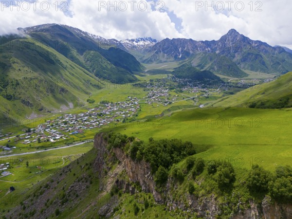 Lush valley with small village and rock face, framed by forested mountains under clouds, aerial