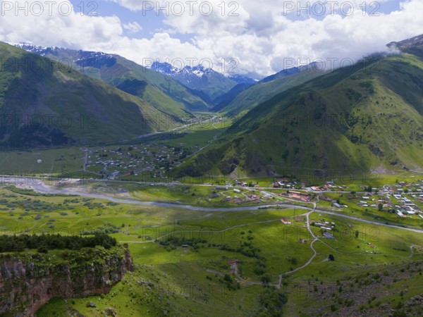 Green valley with river and village, surrounded by high mountains and cloudy sky, aerial view, view