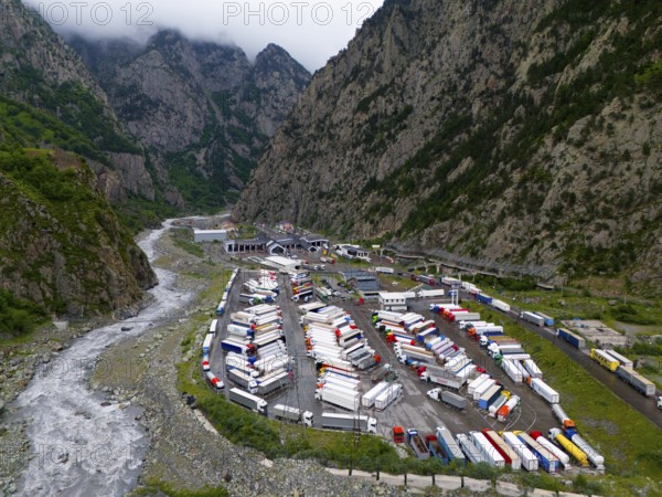 Car park full of lorries in a narrow mountain valley next to a river under a cloudy sky, aerial