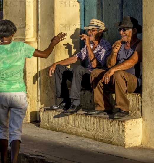 Two old Cubans smoke cigars and sit on a staircase in the warm light of the sunset, they greet a