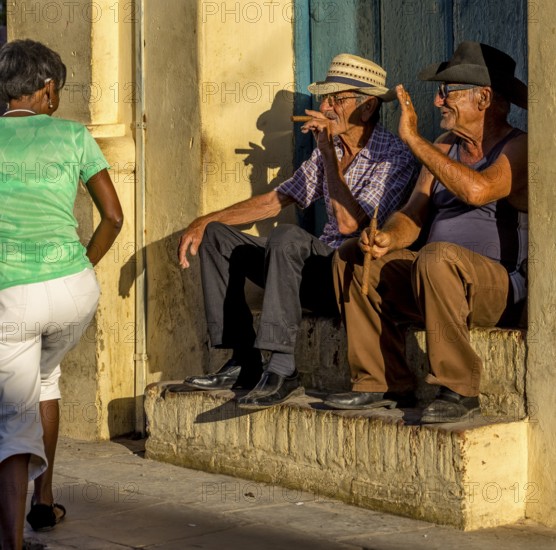 Two old Cubans smoke cigars and sit on a staircase in the warm light of the sunset, they greet a