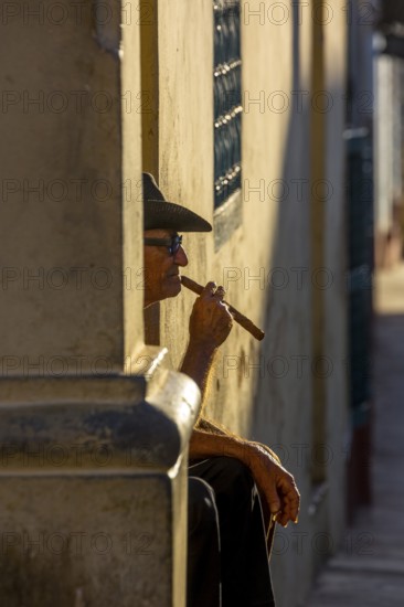 An old Cuban smokes a cigar and sits on a staircase in the warm light of the sunset, Trinidad,