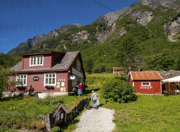 Tourists at mountain hut station, red wooden houses, gravel path, mountains, meadow, blue sky,