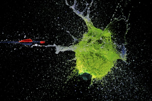 A green water balloon bursts powerfully under the impact of an arrow, against a dark background,
