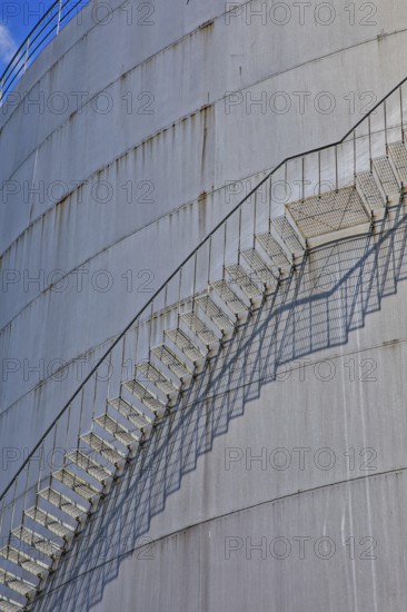 A long metal ladder stretches along a large cylinder building with shadows in the background,