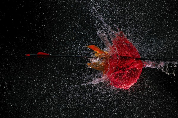 An arrow pierces a red water balloon and creates an impressive water explosion against a dark