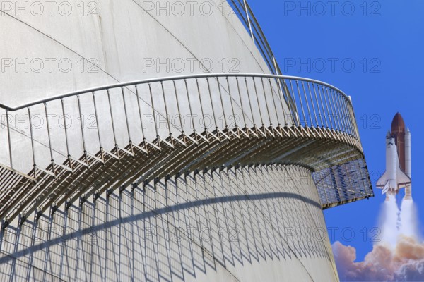 A rocket launches next to a large tank with a metal staircase in the foreground, elevated tank,