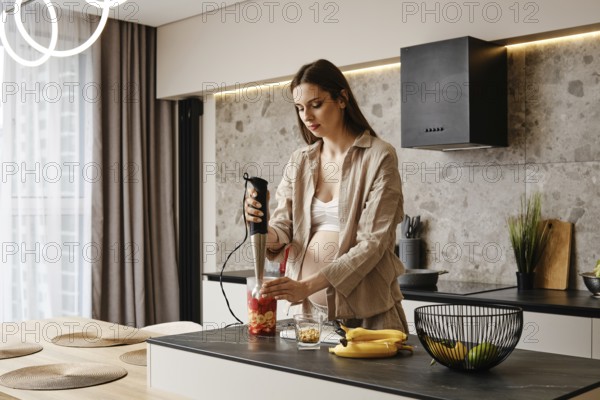 In a stylish kitchen, a pregnant woman blends fresh fruits like bananas and strawberries. Sunlight