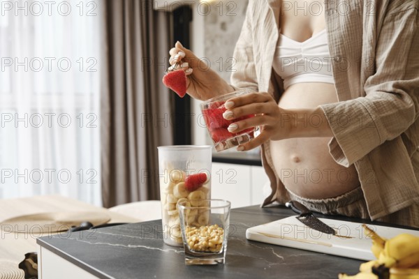 A pregnant woman is creating a nutritious smoothie in a stylish kitchen. She layers strawberries