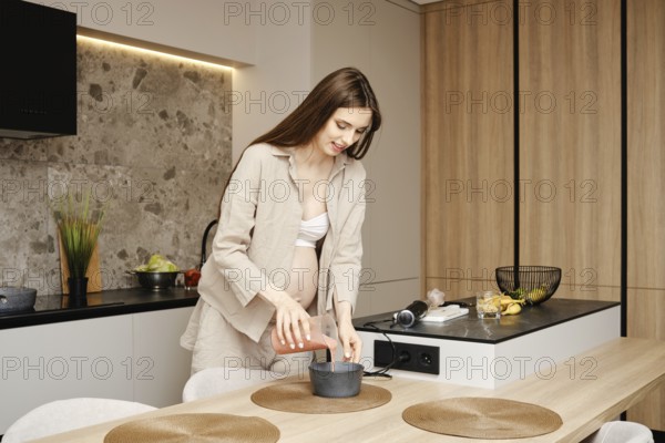 A woman in early pregnancy mixes ingredients in a pot while standing in her stylish kitchen. She