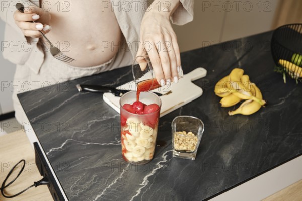 A person pours strawberry syrup over a mix of fruits, including bananas and strawberry, in a glass.