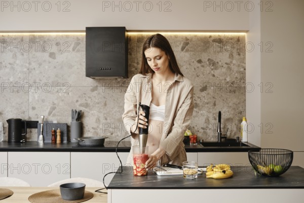 A pregnant woman stands in a contemporary kitchen using a handheld blender to prepare a fruit