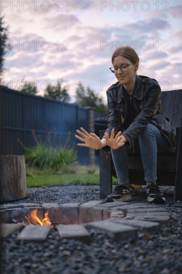 A young woman sits on an outdoor chair near a fire pit, warming her hands in the glow of the flames