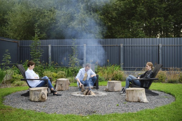A family sits comfortably on wooden chairs, gathering around a fire pit in a spacious garden. Smoke
