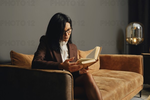A person with glasses is deeply engrossed in reading a book while seated on a stylish couch in a