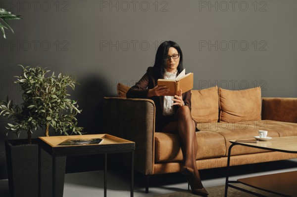 A woman in professional attire sits comfortably on a brown sofa, reading a book intently. A small