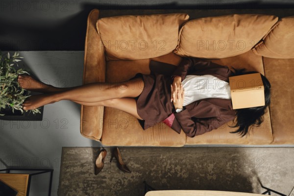 A woman lounges comfortably on a stylish couch, dressed in a suit. She places a book over her face,