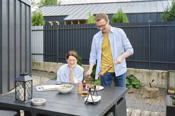 Middle-aged couple enjoy a barbecue meal outdoors in a backyard. Man is serving skewers on a grill,